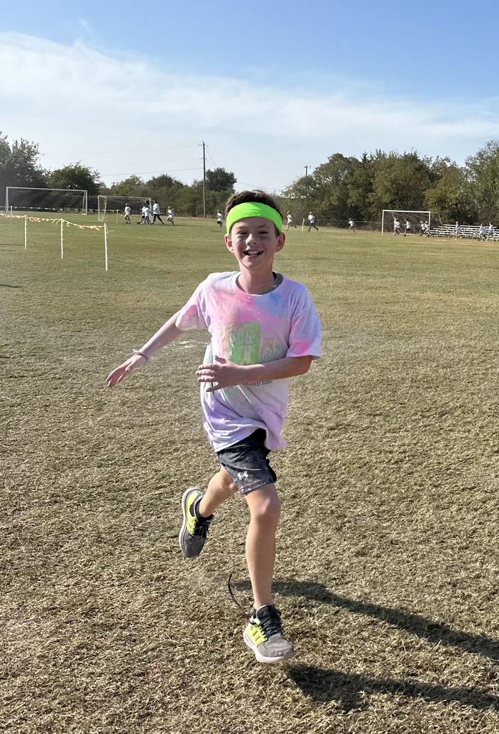 A smiling student in a neon green headband and a color-powder-covered Jogathon shirt running across the field