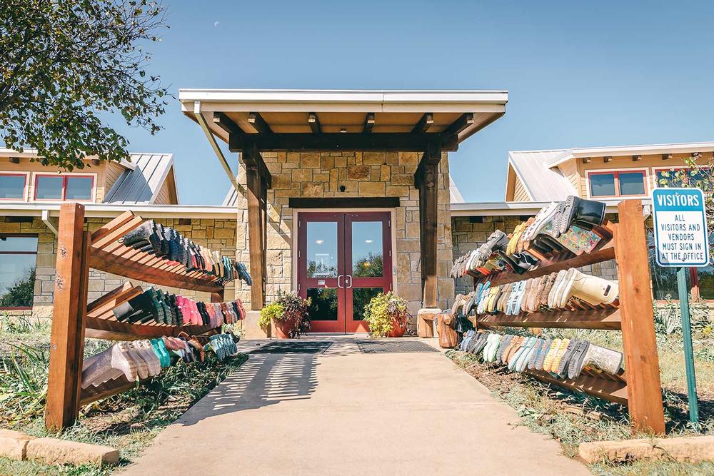 The front entrance of Terra Verde Discovery School, a stone building with red doors, with wooden racks of colorful kids' rain boots lining the walkway